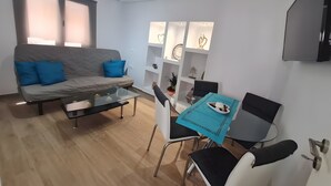 A modern living room with a grey sofa, blue cushions, a glass coffee table, a dining table with chairs, and a TV mounted on the wall. - Maria's Heaven House - Old Town Apartment with Private Terrace, Wi-Fi & Air Conditioning (Rodos)