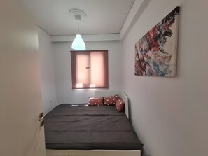 Ein Schlafzimmer mit einem Bett, einem Fenster mit Jalousien, einer Deckenleuchte und einem an der Wand hängenden abstrakten Gemälde.