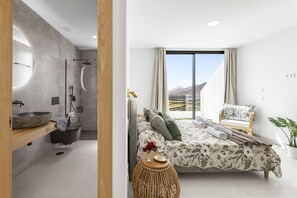 A modern bathroom with a large basin, shower, and a wooden shelf. The bedroom features a bed with a floral bedspread, a view of the mountains, and a wicker side table. - Villa Infinity Views 3 by Escape Home (Tahiche)
