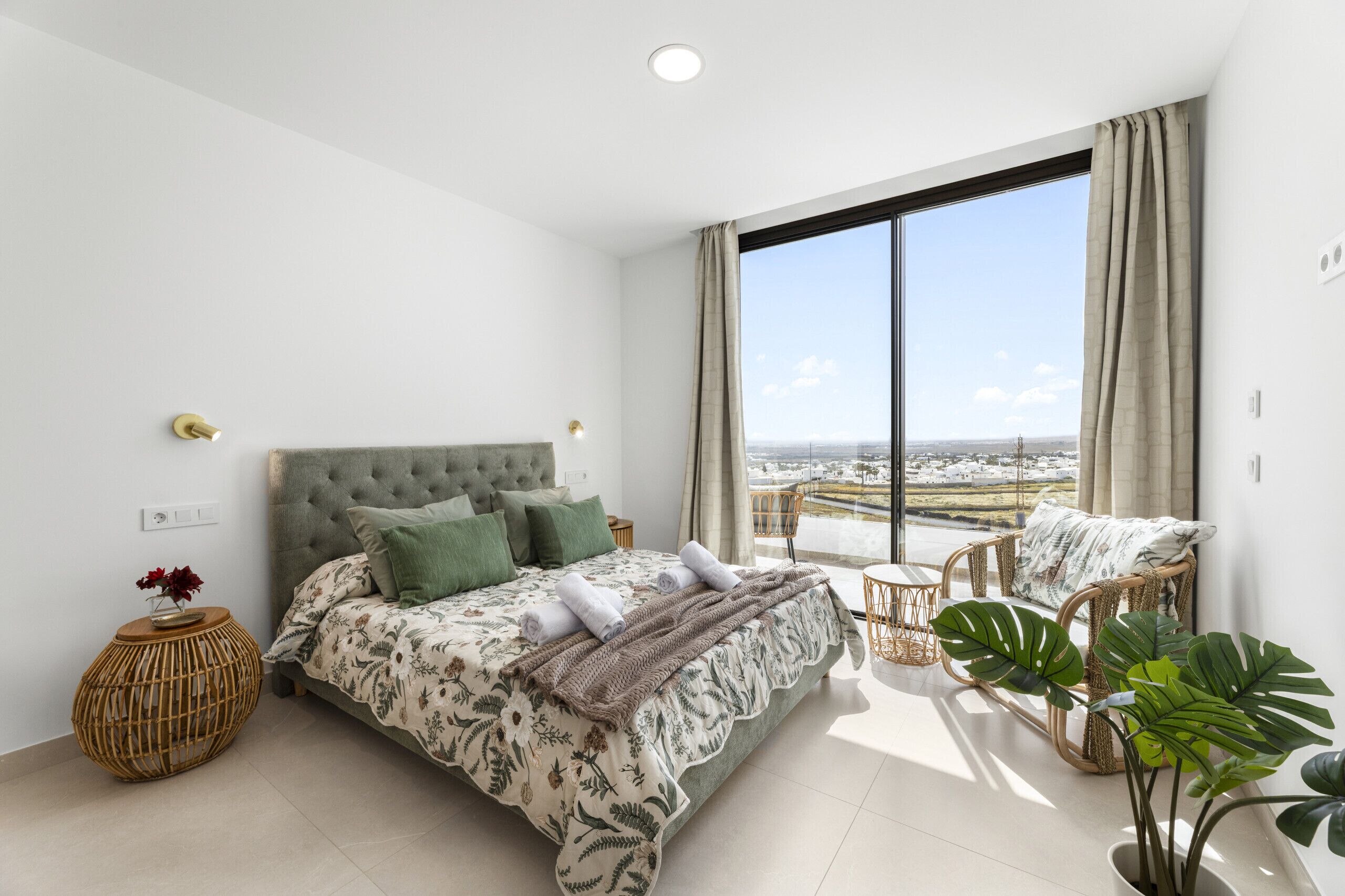 A bedroom with a large bed, a view of the city, and a balcony with wicker furniture.