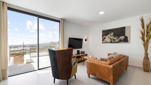 A modern living room with a large sliding glass door, a flat-screen TV, a sofa, a chair, and a painting on the wall. - Villa Infinity Views 4 by Escape Home (Tahiche)