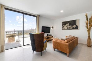 A modern living room with a large sliding glass door, a flat-screen TV, a sofa, a chair, and a painting on the wall. - Villa Infinity Views 4 by Escape Home (Tahiche)