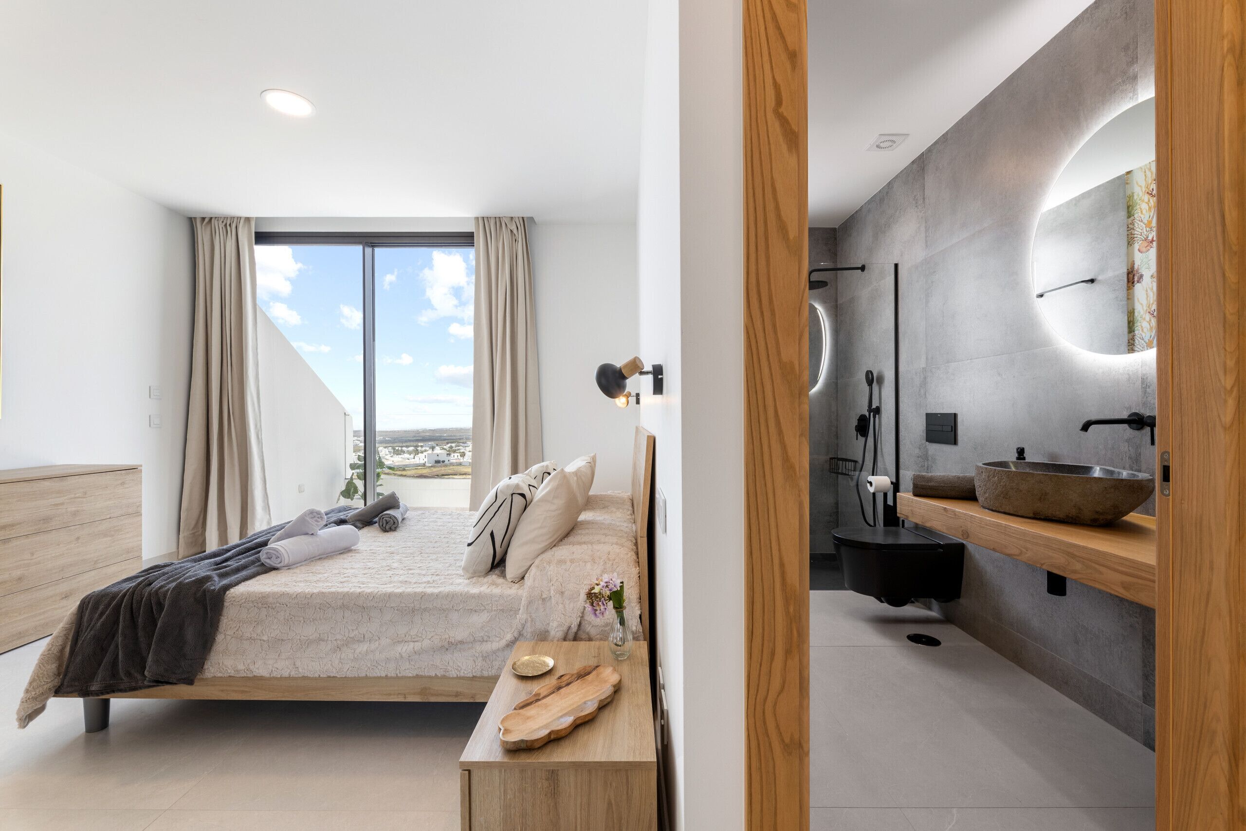 Une chambre moderne avec un grand lit, une table de chevet en bois, une commode, une salle de bain avec un grand miroir, un lavabo et une douche.