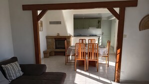 A living room with a sofa, a fireplace, a dining table with chairs, and a kitchen in the background.
