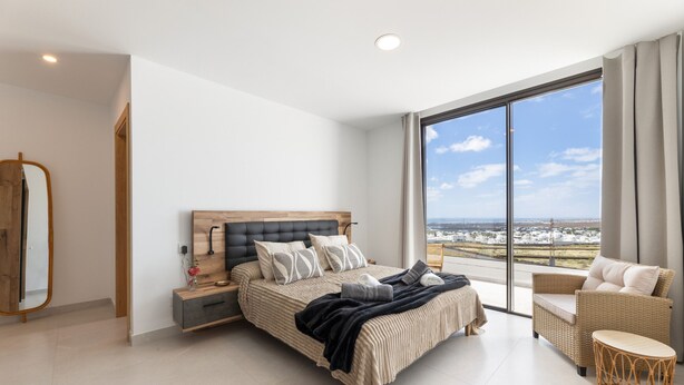 Un dormitorio moderno con una cama grande, una mesita de noche, una silla y vistas a la ciudad a través de amplios ventanales.