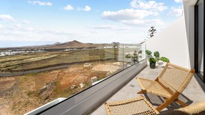 Un balcón con mobiliario de mimbre, barandilla de vidrio y vista a un paisaje con campos y montañas al lejano.