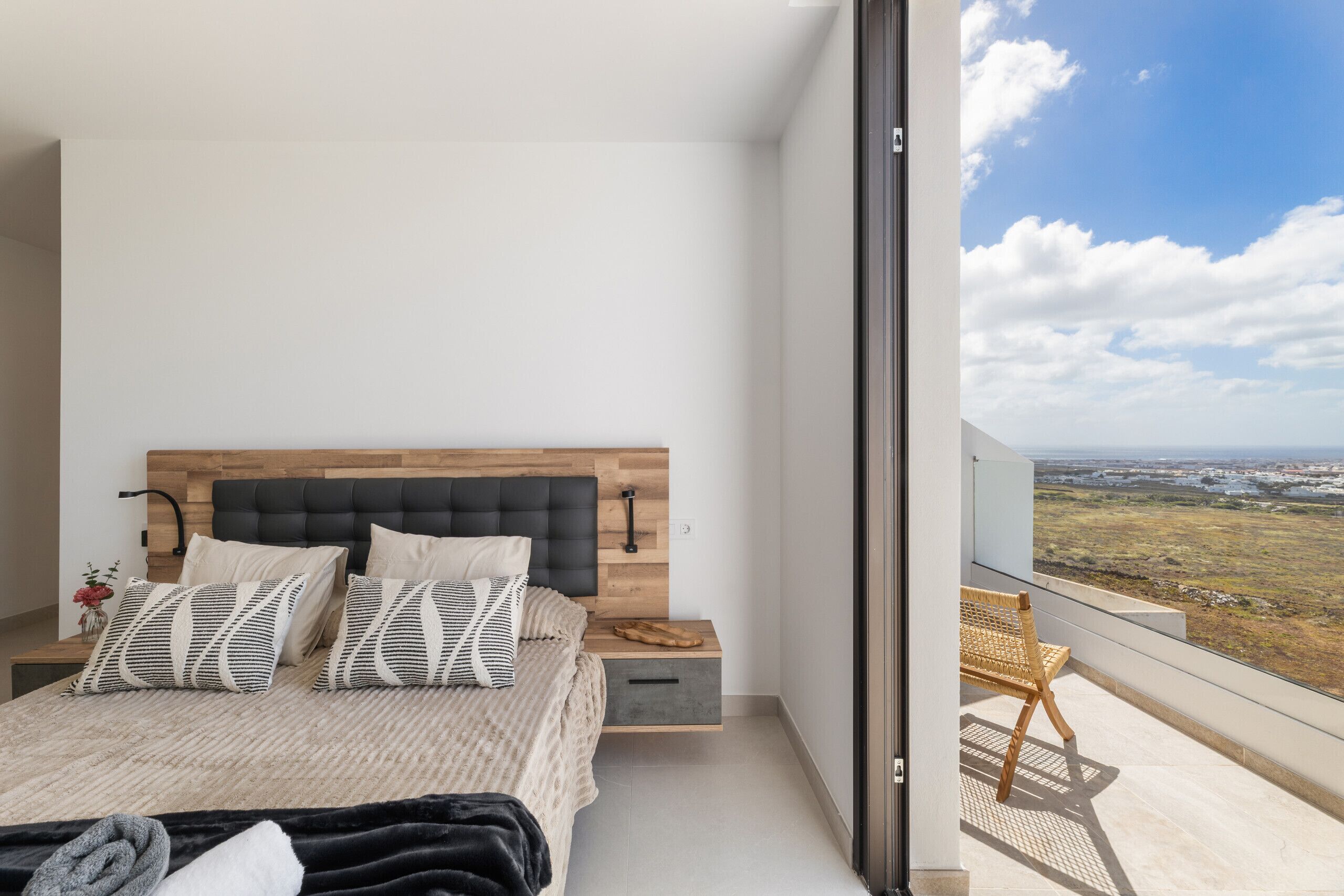 Una camera da letto moderna con una testiera in legno, un letto grande con cuscini decorati, un comodino e vista su un balcone con una sedia in vimini.