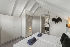 Ein modernes Schlafzimmer mit schrägen Wänden, einem Bett mit weißen Leinen, einem dunklen Kopfteil und einer Wandleuchte.