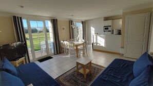 A living room with a TV, blue sofas, a dining table with chairs, a kitchen area, and large windows with curtains. - House Port Bourgenay 6-8 pers - 3 bdrms - pools/toboggans-Plage-Golf- Activities (Talmont-Saint-Hilaire)