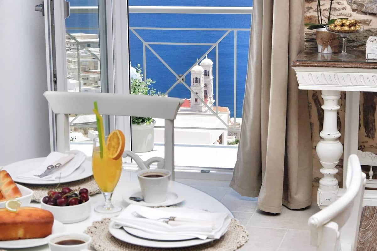 A dining area with a table set for breakfast, a view of a blue sea, a white building, and a stone wall.