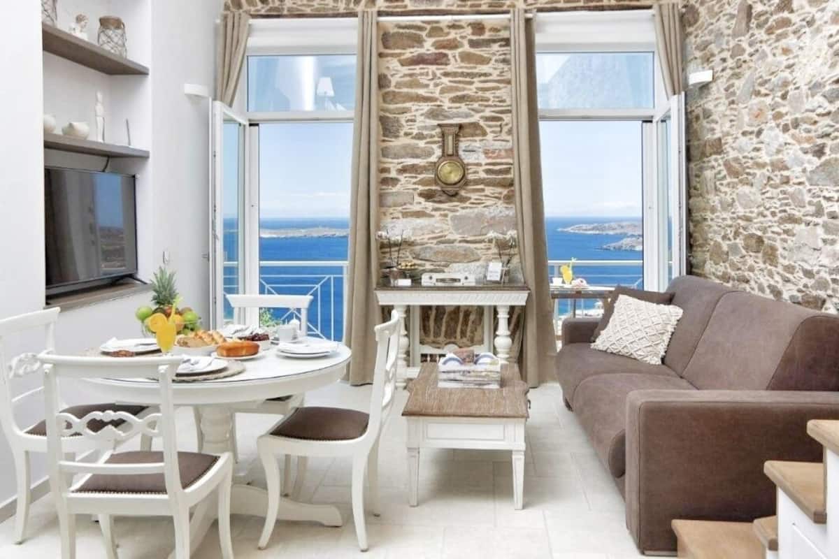 A room with a stone wall, a dining table with chairs, a sofa, and a view of the sea.