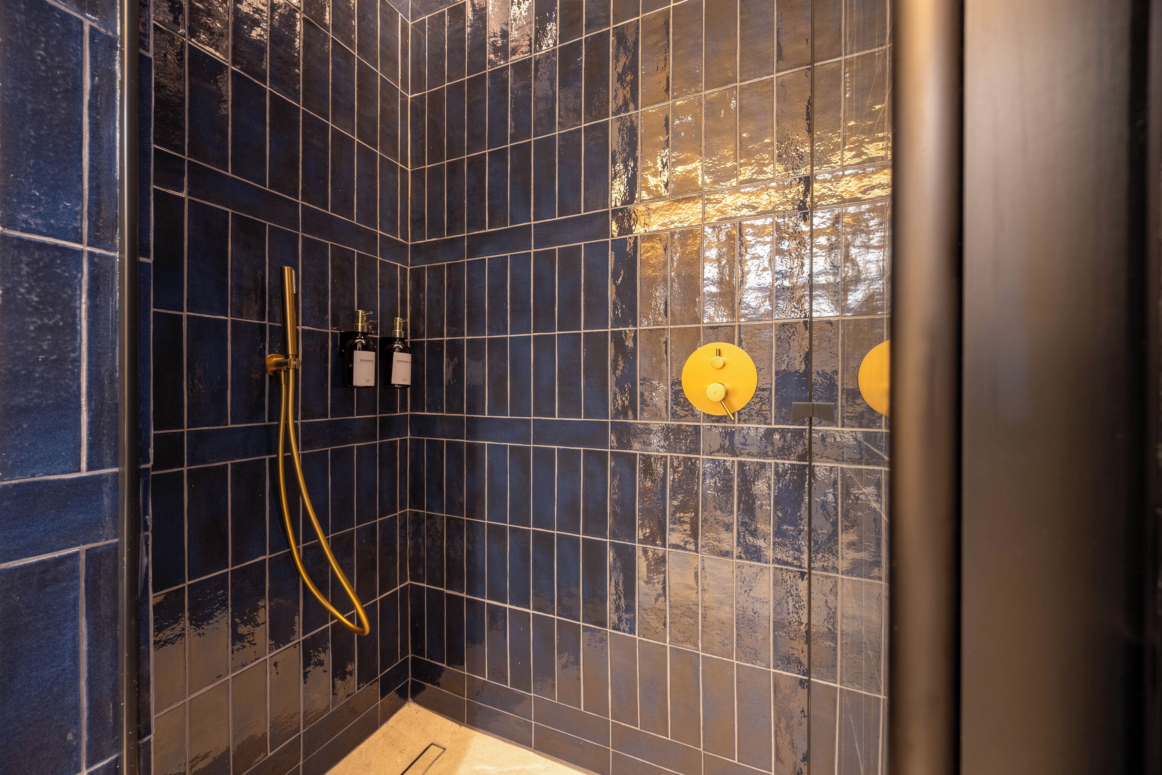 Een hoek van een ruimte met donkerblauwe betegelde muren, een geel douchekop en een goudkleurige douche slang.
