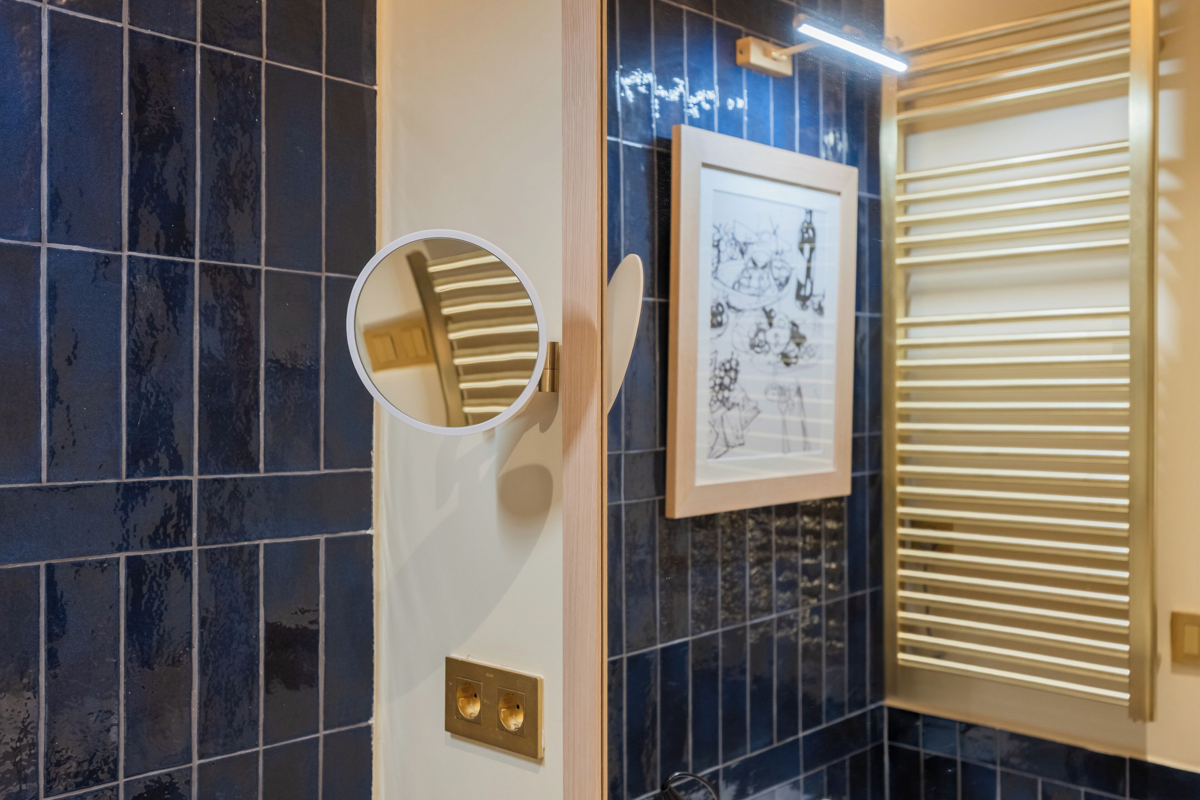 Un baño con azulejos azul oscuro, un espejo redondo, una obra de arte enmarcada y una ventana con persianas.