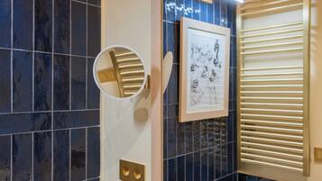 Un baño con azulejos azul oscuro, un espejo redondo, una obra de arte enmarcada y una ventana con persianas.