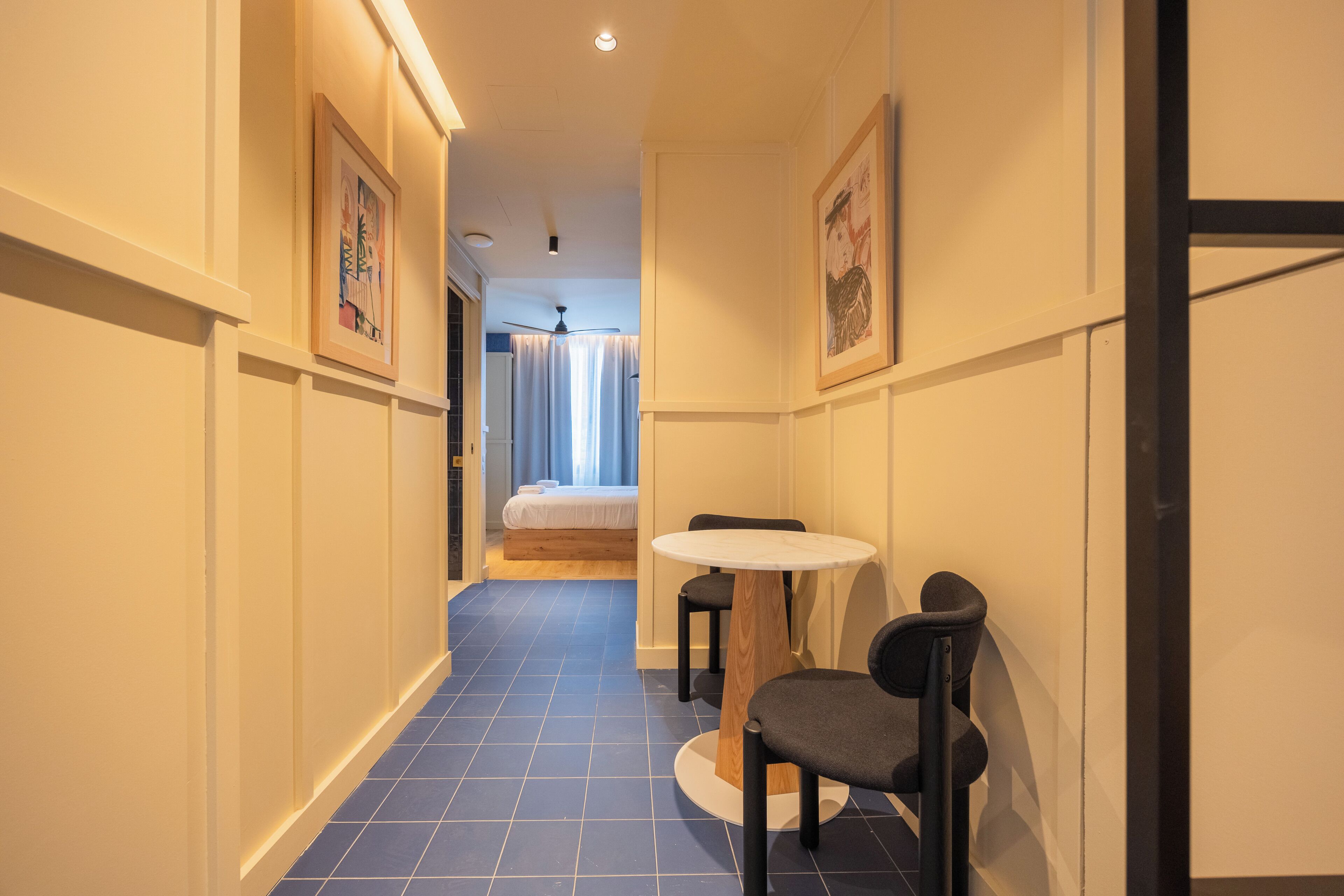 A narrow hallway with a bed, a table with chairs, and framed pictures on the walls.