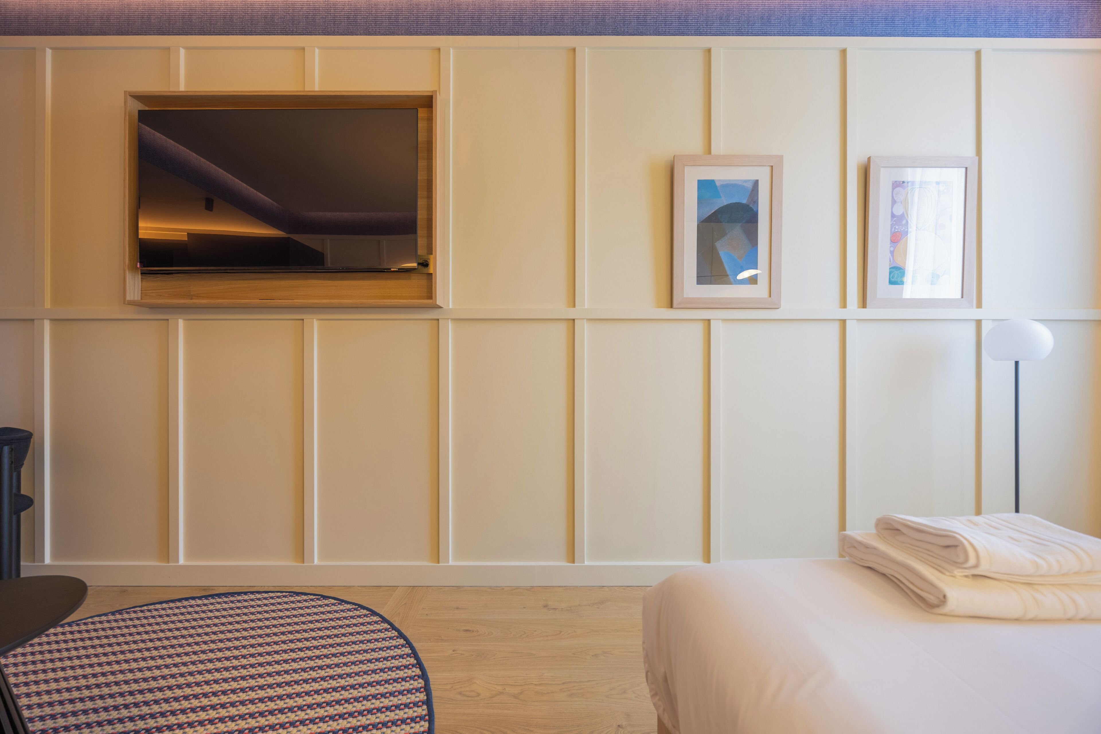 A hotel room with a bed, a desk, a chair, a wall-mounted TV, and framed pictures.