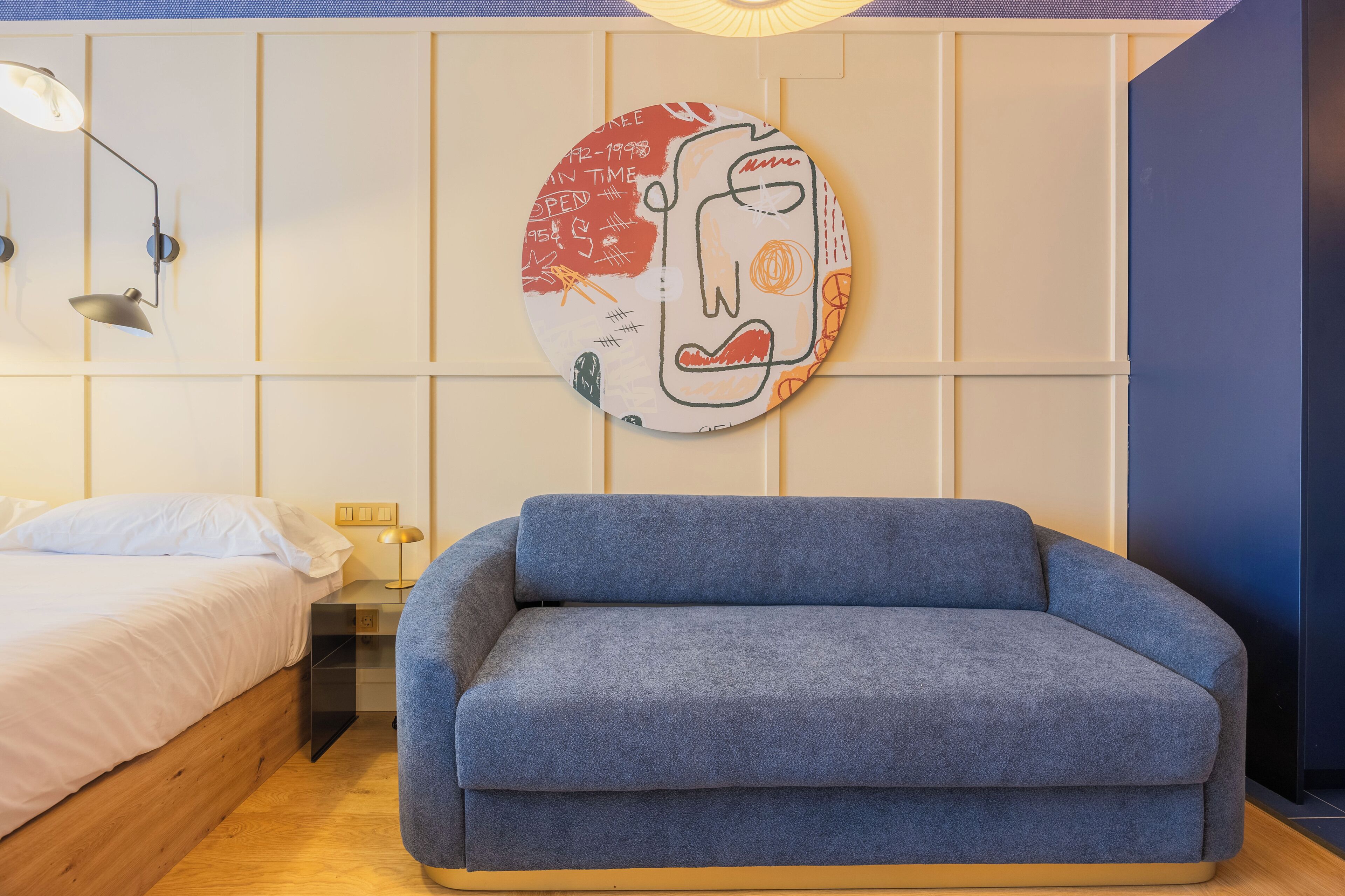 A room with a bed, a blue sofa, a wall-mounted lamp, and a circular artwork.