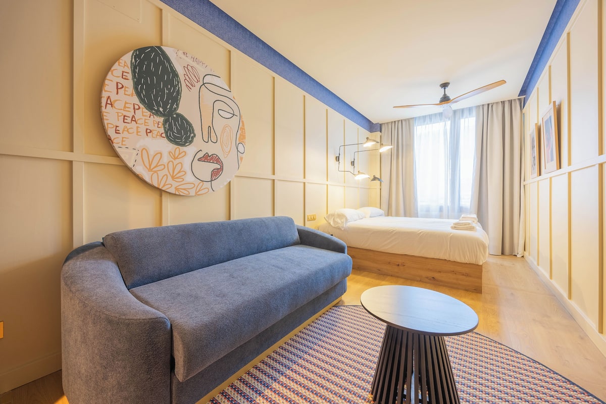 A modern hotel room with a sofa, a round table, a bed, and a wall with a circular artwork.