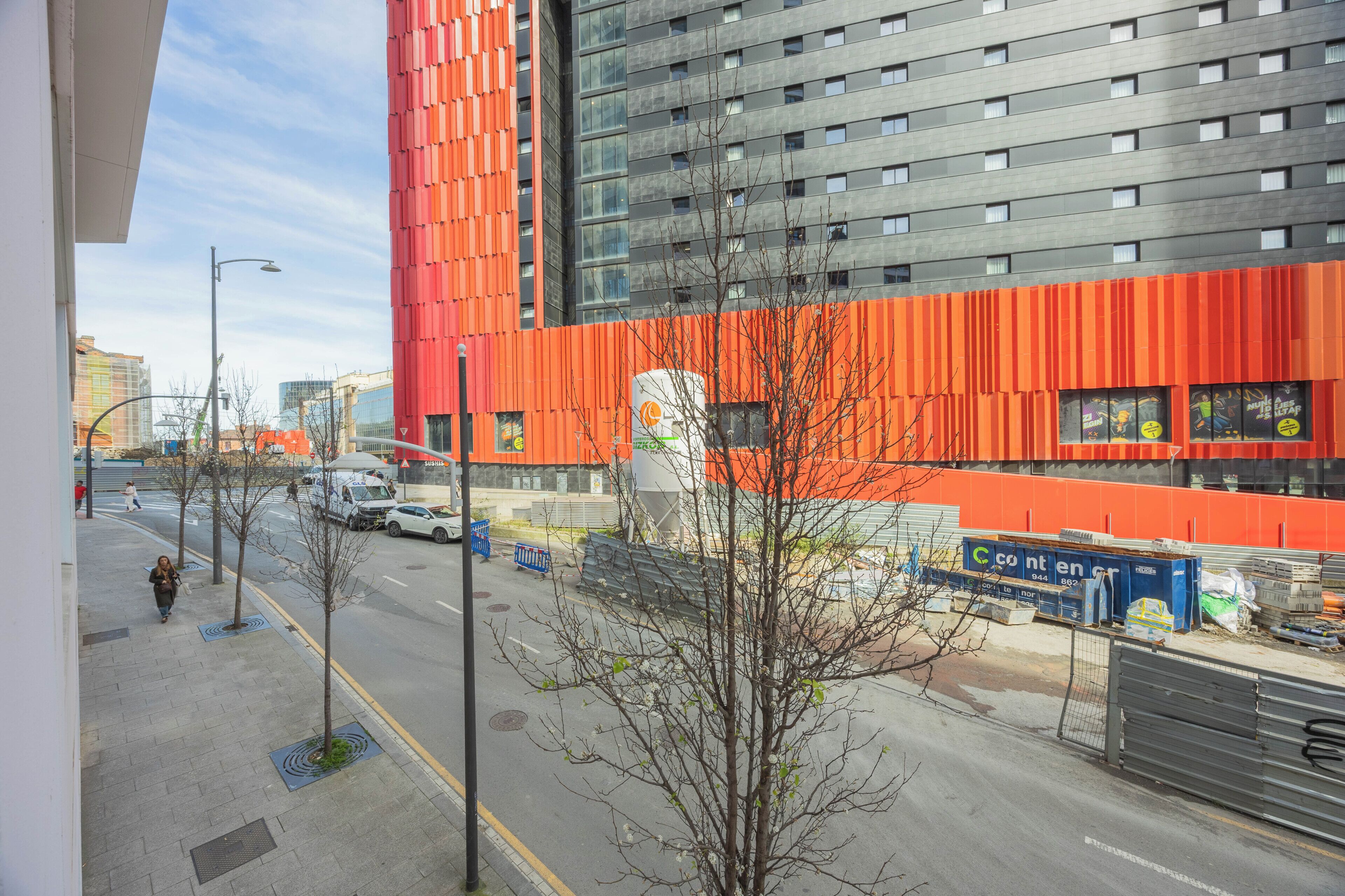 Un bâtiment moderne à la façade rouge, avec une promenade piétonne et des voitures garées.
