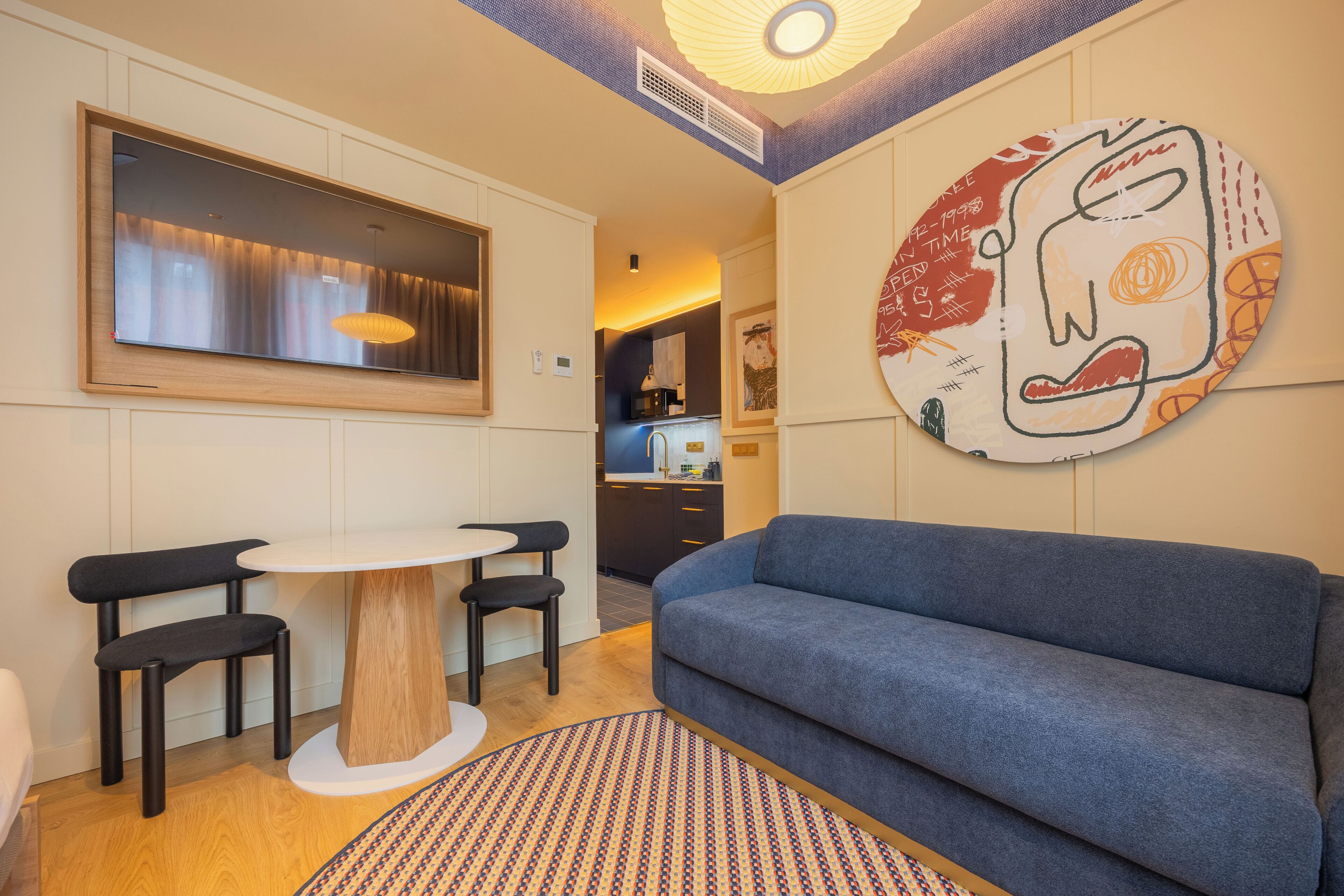 Un salon moderne avec un canapé, une table ronde entourée de chaises, une grande œuvre d’art accrochée au mur et une cuisine en arrière-plan.