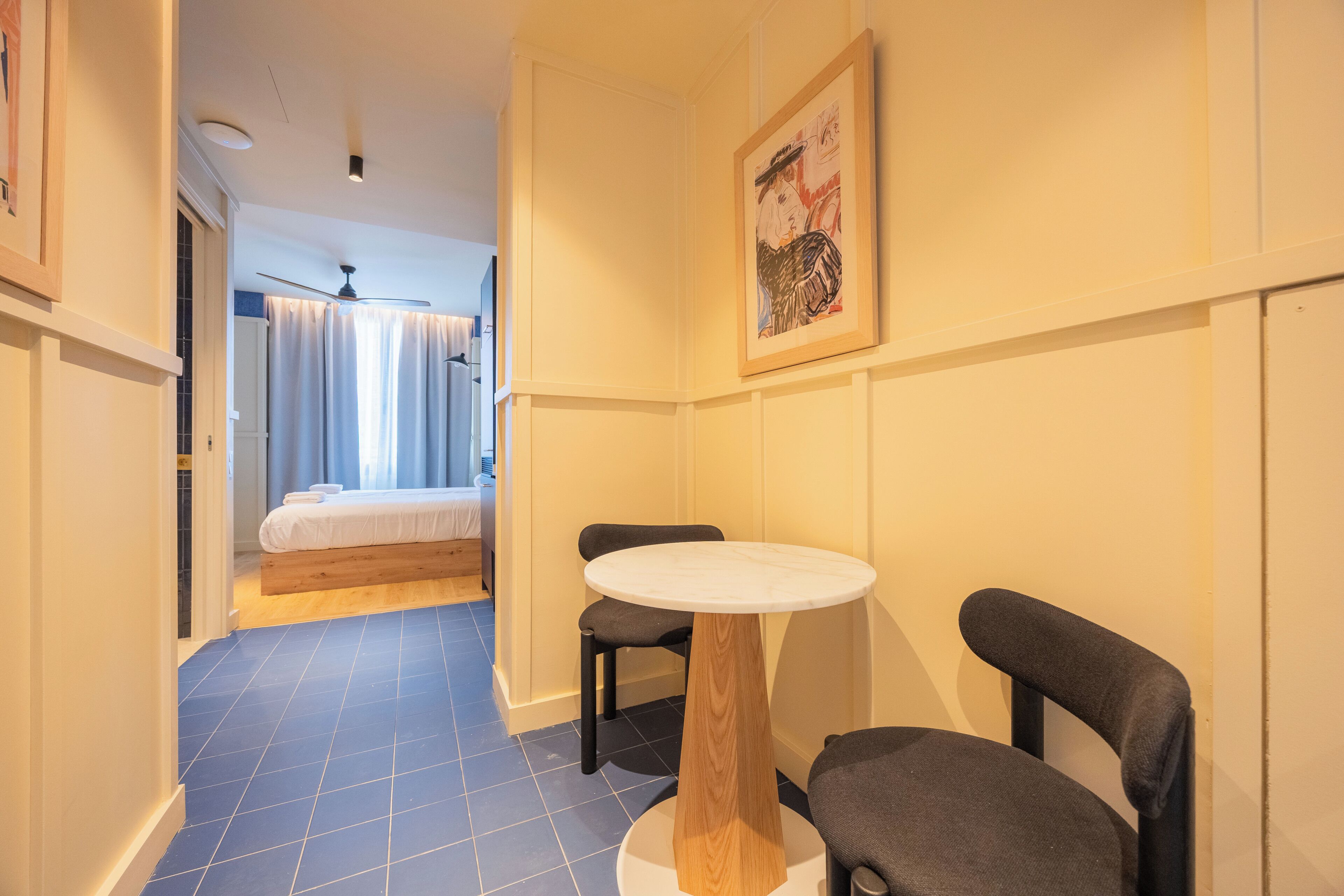 Una habitación de hotel con una cama, una mesa redonda con sillas, un ventilador de techo y un cuadro en la pared.