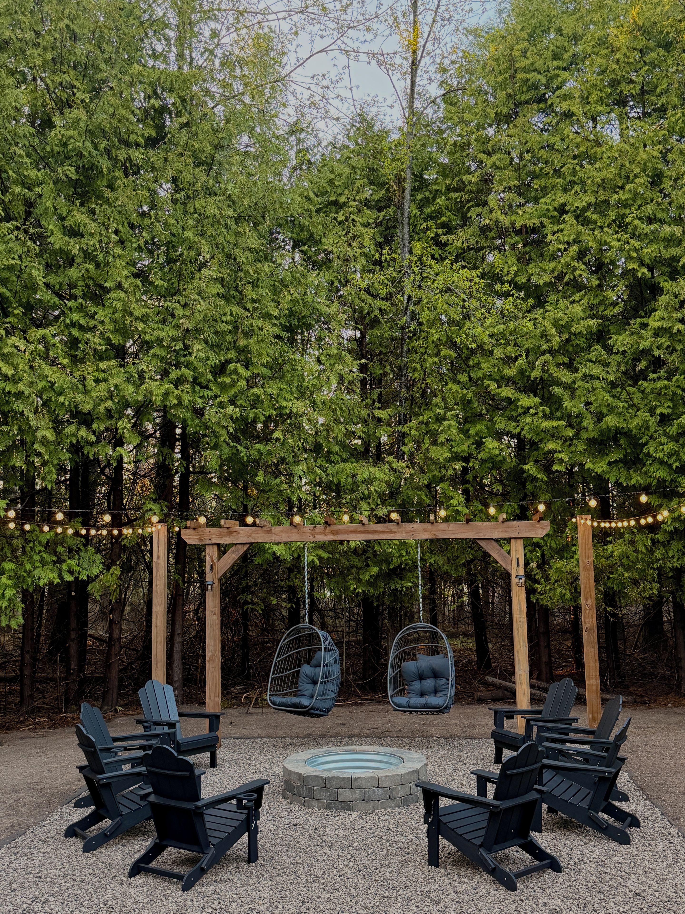 A wooden pergola with hanging chairs and a fire pit surrounded by gravel, set in a forested area.