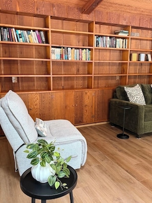 A cozy living room with a wooden bookshelf, a grey armchair, a green sofa, and a small table with a plant. - 4BR house in Cheboygan with a game room and a private boat slip nearby (Cheboygan)