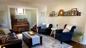 Ein Wohnzimmer mit einem Klavier, einem blauen Sessel, einem dunkel ledernen Sofa und einem gemusterten Teppich.