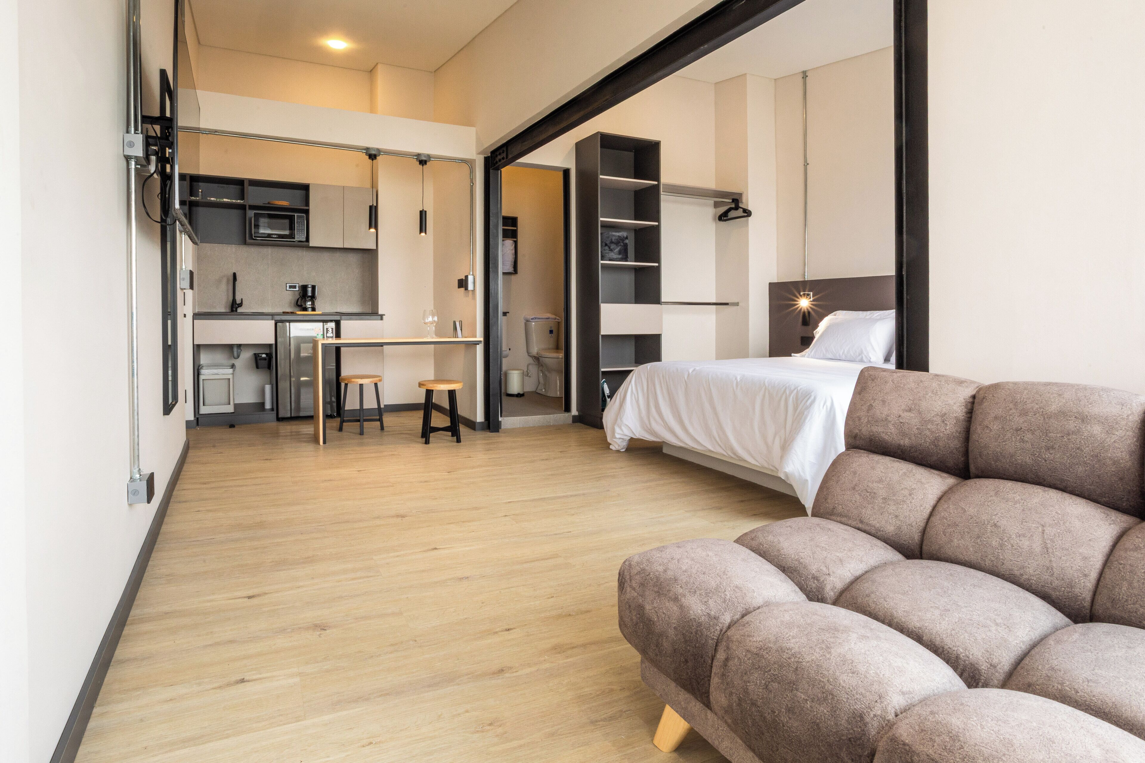 A modern hotel room with a sofa, a bed, a kitchenette, and a bathroom.