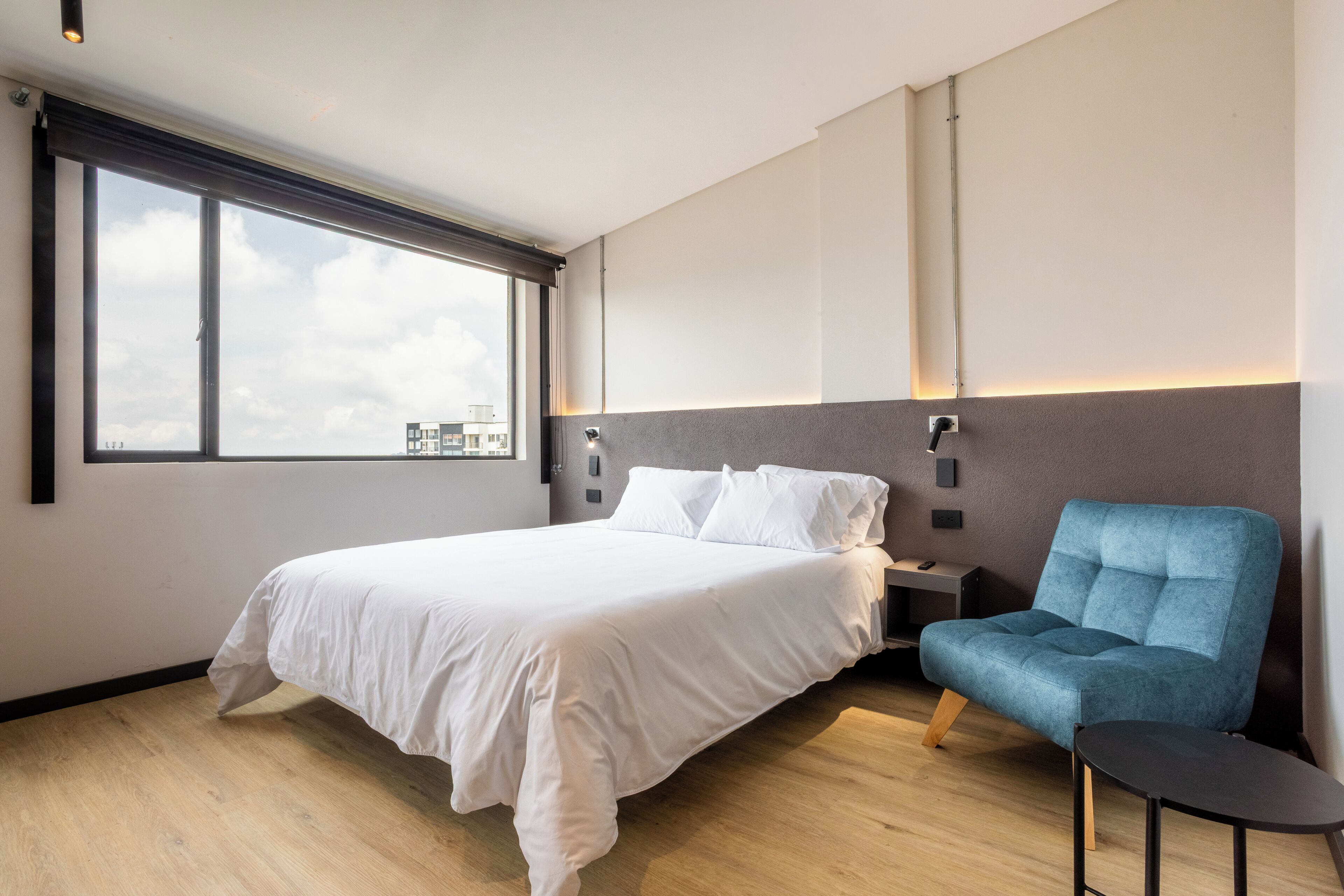 Un hotel moderno con una cama grande, un sillón azul, una mesita y vistas a la ciudad desde la ventana.