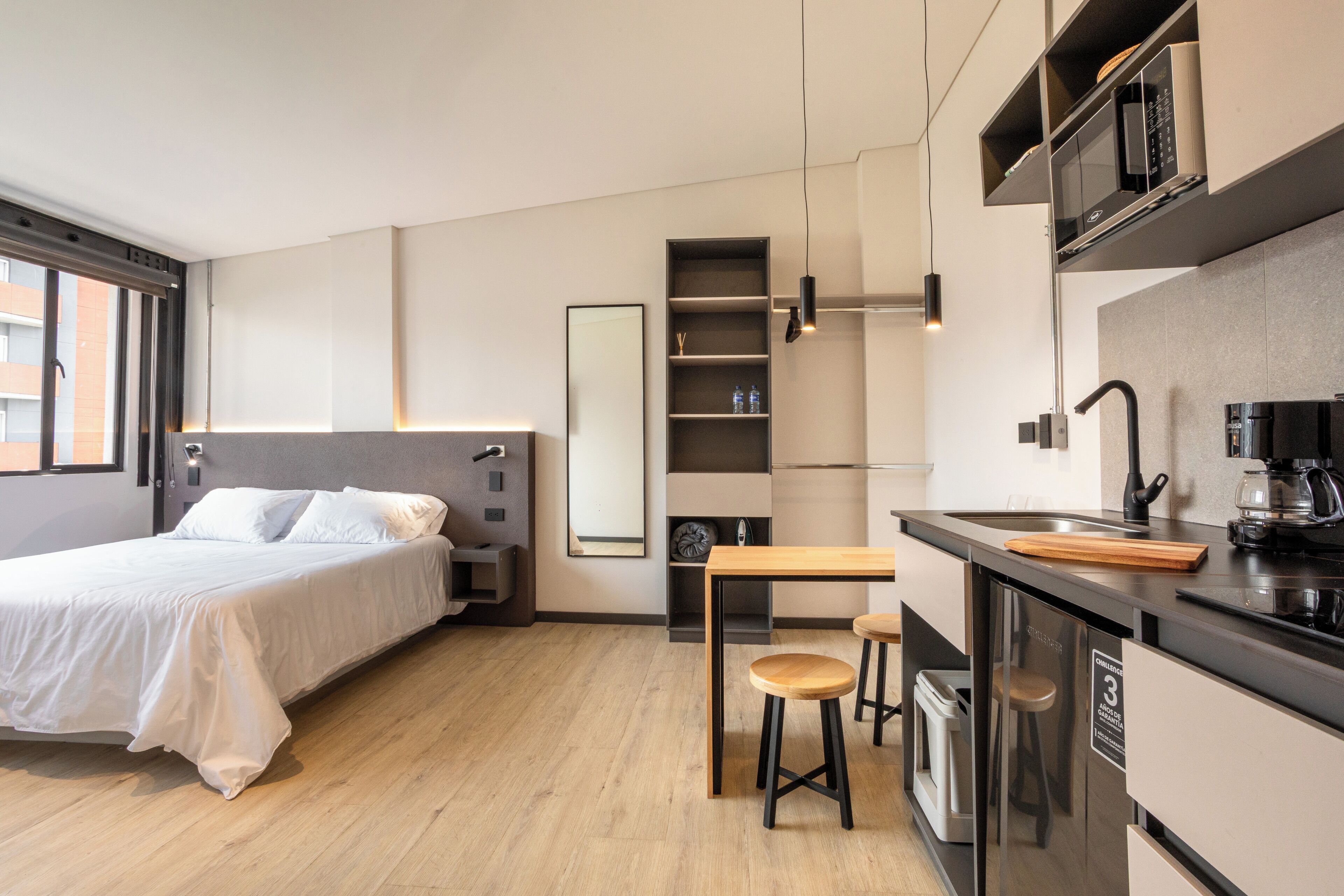 A modern hotel room with a bed, a small kitchenette, a dining area with stools, and a large window.