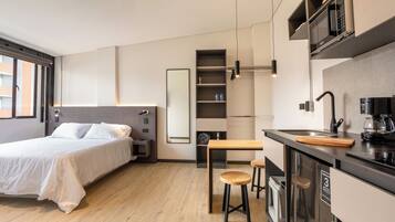 A modern hotel room with a bed, a small kitchenette, a dining area with stools, and a large window.