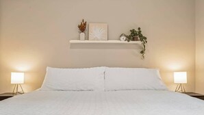 Un lit bien fait, avec une literie blanche, une étagère au-dessus ornée d’objets décoratifs et deux lampes de chevet.