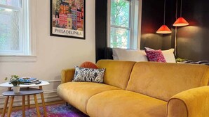 Een gele bank, een kleurrijk vloerkleed, een klein tafeltje met een plant en een lijst met foto aan de muur.