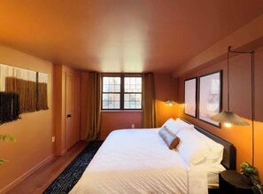 Una camera d'albergo con un letto, una finestra, una porta, una lampada a parete e una pianta.