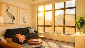 A living room with a black sofa, a round wooden coffee table, a patterned rug, and a large window with a view of buildings.