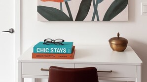 A white desk with a framed floral artwork, a stack of books, a pair of glasses, and a brass container.