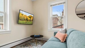 Un salon avec un canapé, un téléviseur fixé au mur, une petite table ronde et une fenêtre donnant sur des immeubles.