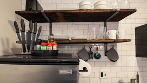 Une cuisine équipée d’un réfrigérateur en acier inoxydable, d’une étagère en bois où sont rangés de la vaisselle, et d’ustensiles de cuisine suspendus.