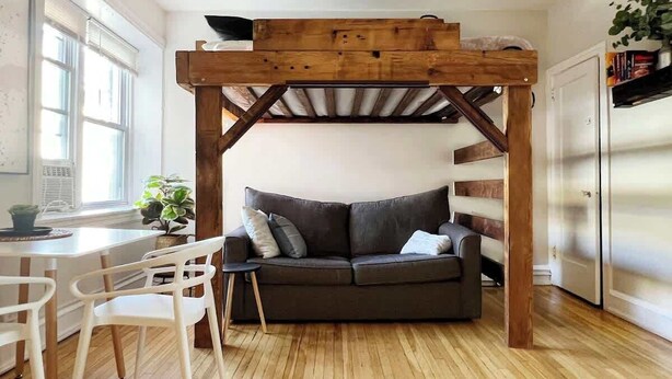 A cozy room with a wooden ceiling structure, a grey sofa, a white chair, and a small table with a plant. - FIFA Walkable King Bed 3 Beds (Philadelphia)