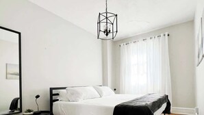A bedroom with a bed, a bedside table, a lamp, a hanging light fixture, and a window with curtains. - FIFA Walkable King Bed 3 Beds (Philadelphia)