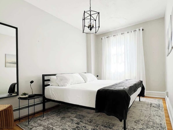 A bedroom with a bed, a bedside table, a lamp, a hanging light fixture, and a window with curtains. - FIFA Walkable King Bed 3 Beds (Philadelphia)