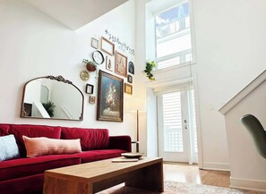 Ein Wohnzimmer mit einem roten Sofa, einem Holztisch und einem großen Spiegel. An der Wand befinden sich gerahmte Bilder, eine Stehlampe und eine Treppe, die in einen oberen Bereich führt.