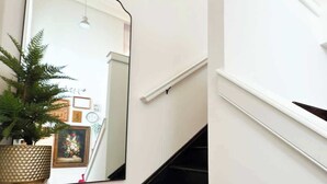 En hall med en stor spegel som reflekterar en trappa, en krukväxt och en inramad tavla på väggen.