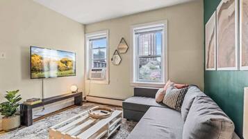A living room with a flat-screen TV, a sofa, a coffee table, and a window with a city view.