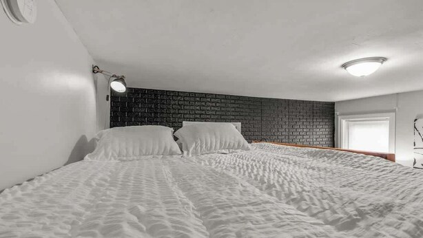 A bedroom with a brick-patterned headboard, a bed with white bedding, a bedside lamp, and a window with blinds. - Huge Desk - Central - Soak Tub - Walkscore 98 (Philadelphia)