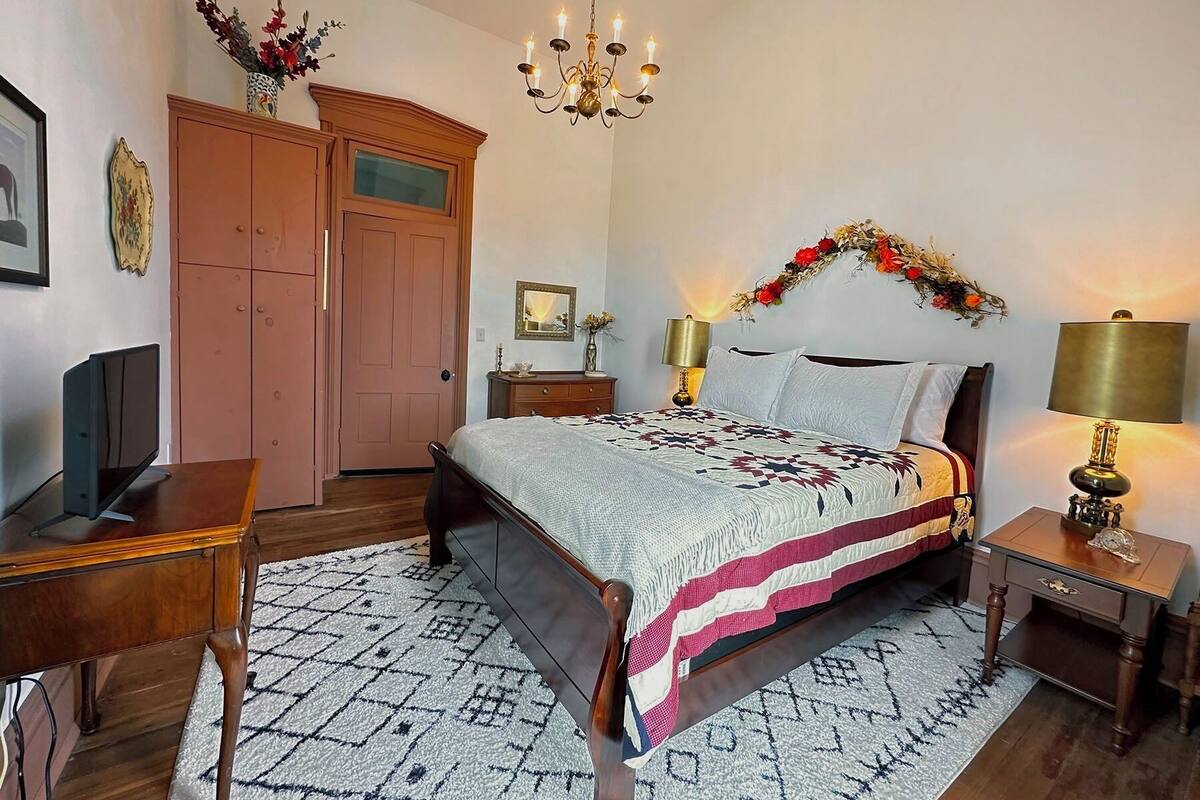 A bedroom with a bed, bedside tables, a desk, a wardrobe, and a chandelier.