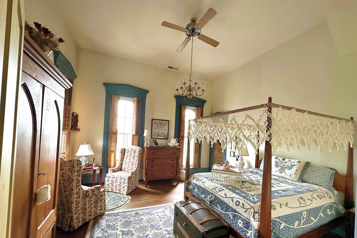 A bedroom with a four-poster bed, a wooden wardrobe, a chair, a dresser, and a chandelier.