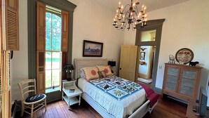 A bedroom with a bed, a chandelier, a window with shutters, a chair, a nightstand, a dresser, and a door. - Rising Hall B&B (Coatesville)
