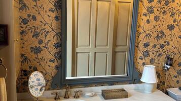 Uma casa de banho com um espelho grande, uma pia, toucador com gavetas, um pequeno banquinho e um painel decorativo com papel de parede floral.